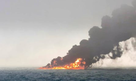 The sea is in the forefront with a strong fire and thick black smoke coming from the vessel