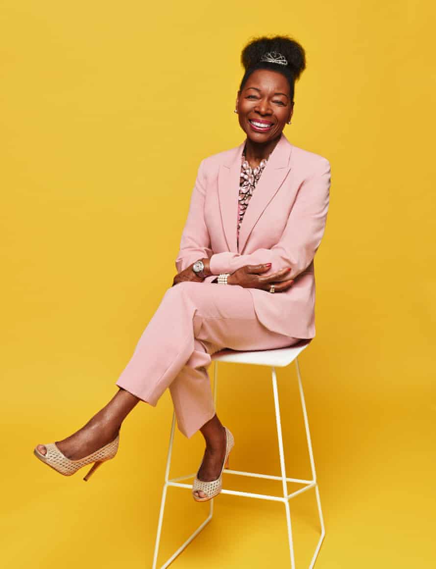 Floella Benjamin in a pink trouser suit sitting on a high stool with her legs crossed, yellow background