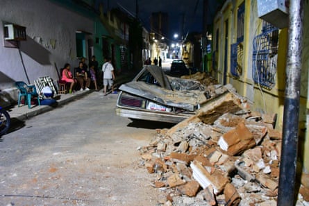 People sit or stand near a damaged car and a pile of bricks and rubble