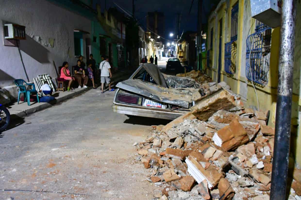 People sit or stand near a damaged car and a pile of bricks and rubble