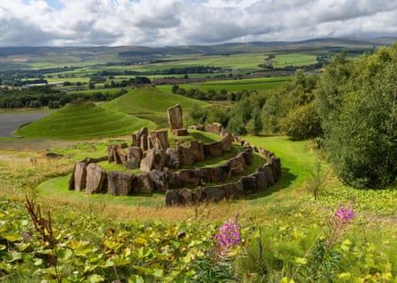 A mound with spiralling stones and a grass pathway in the foreground, and two grass-covered dome-shaped hills behind