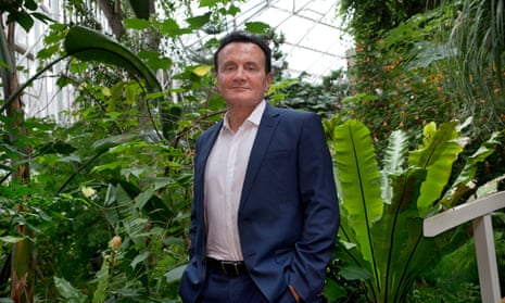 Pascal Soriot in a suit but no tie poses with his hands in his pocket by some lush greenery in a conservatory