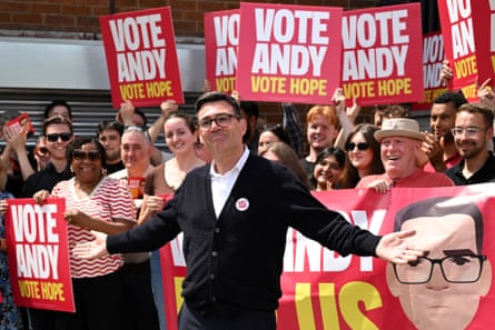 Andy Burnham, wearing a white shirt and black cardigan, stands in front of a small crowd holding up ‘Vote Andy, Vote Hope’ placards