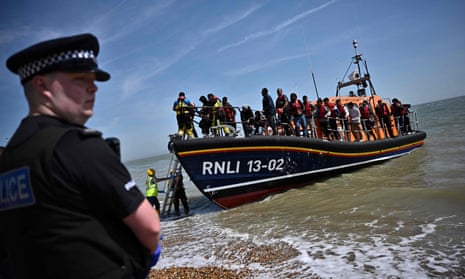 A uniformed police officer stands on the beach as people wearing lifejackets disembark from a lifeboat