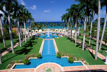 A view out to the ocean of lush green lawns and large blue pools bordered by rows of palm trees, on a sunny day