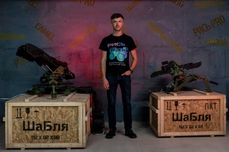 A man in jeans and T-shirt stands between two crates with military machines on top of them