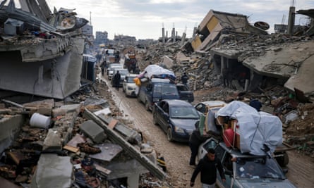 A line of vehicles packed with belongings travel between devastated buildings.