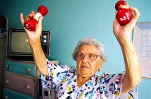 an older woman in a floral dress grimacing slightly as she lifts two small dumbbells