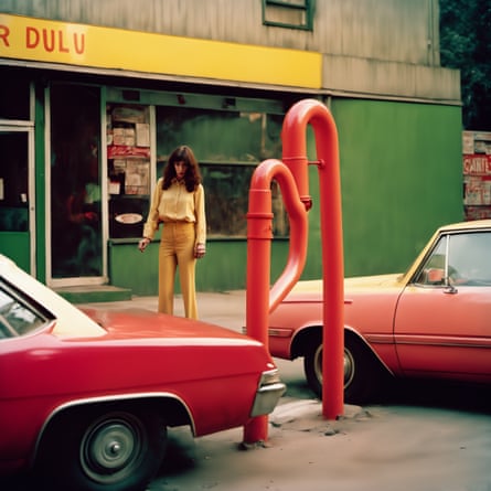 A woman in a bright yellow blouse and orange trousers walking on a pavement past a bright red car