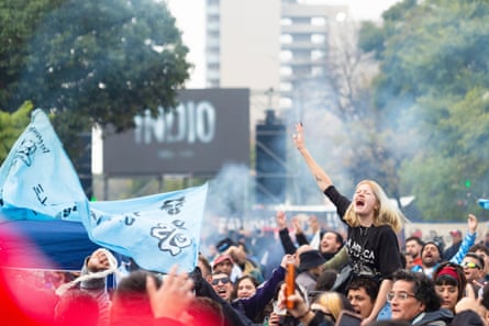 A crowd gathers in a park by a stadium. A young woman with blond hair, sitting on a man’s shoulders, raises a hand and cheers or sings. People are holding banners and flags.