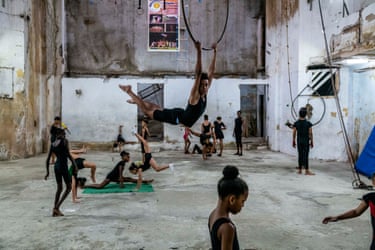 A young circus acrobat swings on a hoop above children training in a deteriorating Havana hall