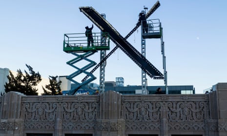 With evening light behind them, two workers in silhouette standing in hydraulic lift buckets raise their arms to adjust a two-story, black X.