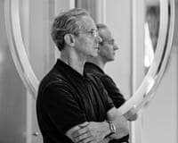 Fabio Luisi in glasses and a black shirt stands with arms crossed in profile, viewed through curved sculptural forms