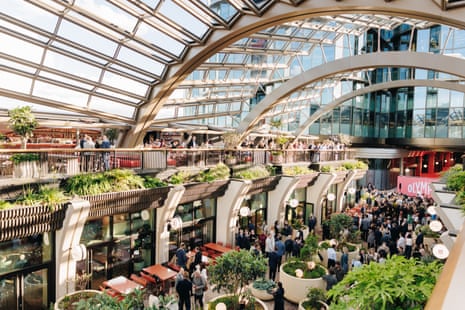 A crowded multi-level indoor venue with curved glass roof, arched beams, planted terraces, and people gathered below