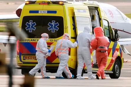 Three figures escort a fourt, all in hazmat suits, to an ambulance