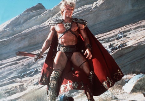 Dolph Lundgren in fantasy warrior costume with shoulder armor and red cape stands on rocky terrain