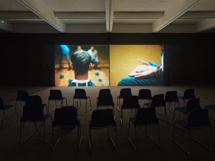 A dark lit room with rows of chairs in front of two video screens, one showing the back of a man’s head, the other a closeup of hands on a lap