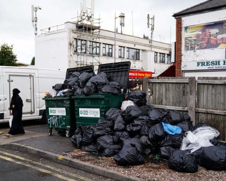 Uncollected refuse bags on the sidewalk and in overflowing bins
