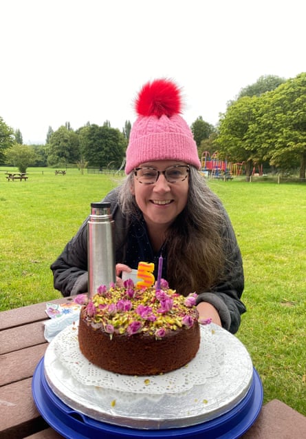 A woman wearing a pink bobble hat with a red pom-pom atop and grey quilted jacket crouches in front of a dark chocolate covered in pistachios and rose petals in a green looking lawn and trees - and possibly a playground in distance on overclouded day