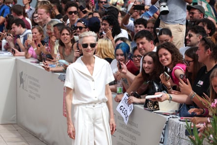 Tilda Swinton wears a white shirt and trousers near a crowd