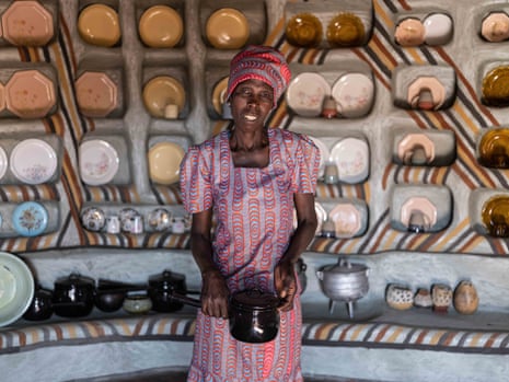 Judith in colourful traditional dress holding cooking pot. Behind her is a wall of plates on decorated shelves