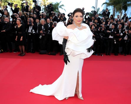 Joan Collins on the red carpet in front of lines of photographers. She wears a long white off-the-shoulder dress with wide sculptural frills and ruffles plus a long train, and long black gloves.