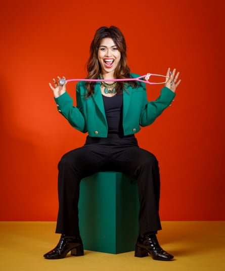 Dr Bhasha Mukherjee sitting on a dark-green cube on a yellow floor, against an orangey-red wall, holding one end of a pink stethoscope in either hand, with her fingers outstretched, wearing a black top, trousers and boots, a short green blazer with gold buttons, and a black and green necklace on a gold chain