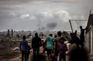 A small crowd of people stand before an expanse of destroyed buildings, watching smoke rising in the distance