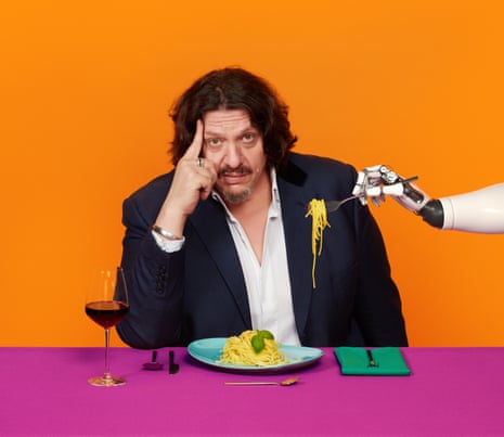 Jay Rayner sitting at a table with a plate of spaghetti in front of him, being offered a fork of food by a robot hand