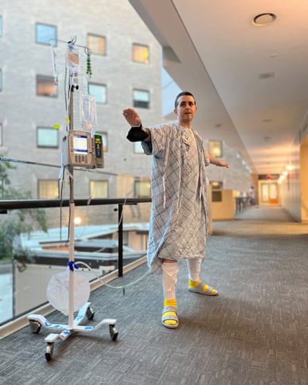 A man connected to an IV drip machine poses in a hospital hallway