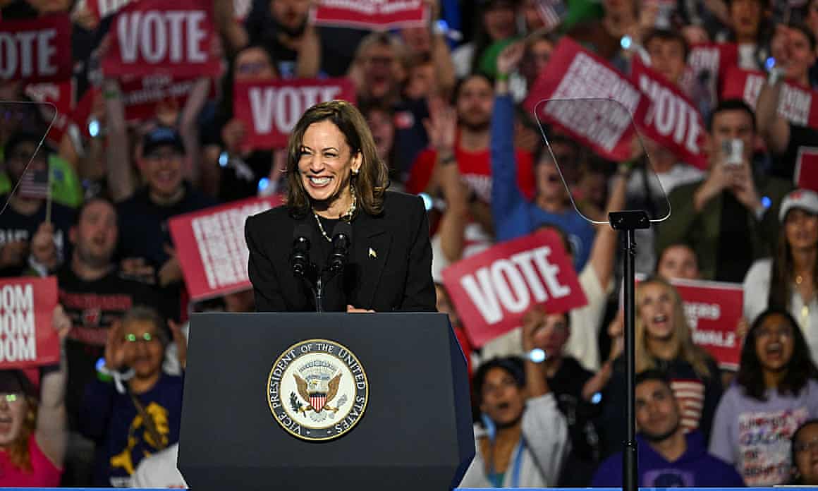Kamala Harris behind a lectern with people waving 'vote' flags in the background
