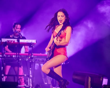 Woman in red body suit and black knee-hight boots plays guitar.
