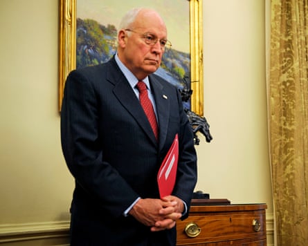 Dick Cheney standing in front of a painting in a yellow-walled room with his hands clasped in front of him holding a red folder
