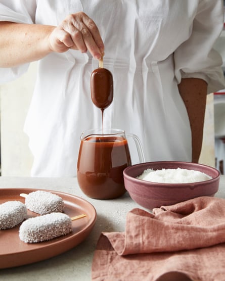 A woman wearing a white shirt dips an ice-cream on a stick into a jug of melted chocolate.