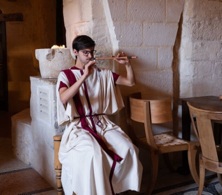 A man wearing red and white robes and glasses, sitting in a stone building playing a wind instrument with two pipes