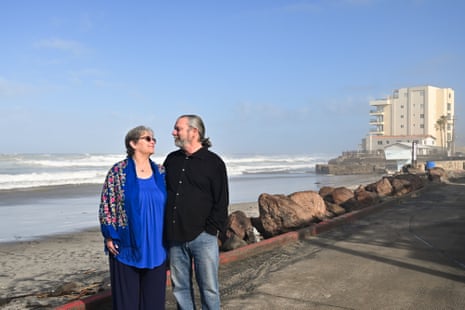 An older white couple gaze at each other, arms around each other, on an asphalt walkway above a beach with a high-rise beyond them.