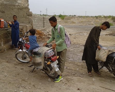 A boy secures ice in panniers on a moped with a smaller child sitting on it in an arid landscape.