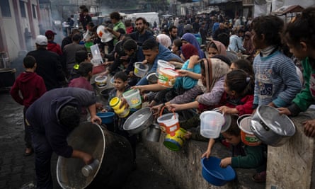 People, mostly women and children, hold empty containers while a worker scrapes a large cauldron of food.