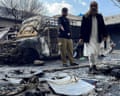 Two men standing in front of burnt-out vehicles look at debris on the ground, including a riot shield