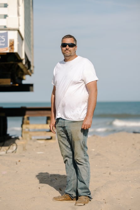 A man in a white T-shirt and blue jeans stands for a portrait on the beach with the ocean as a backdrop