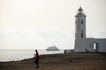 A person walking in front of a lighthouse, with the ship near the horizon in the background