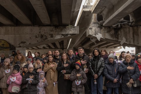 People stand holding their hands to their hearts under the destroyed bridge