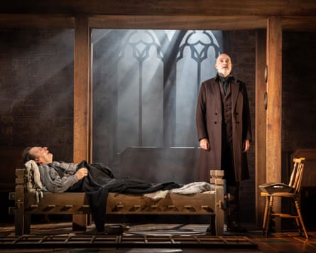 A man lies in bed looking pained while a man in a long brown jacket stands at the end of the bed.