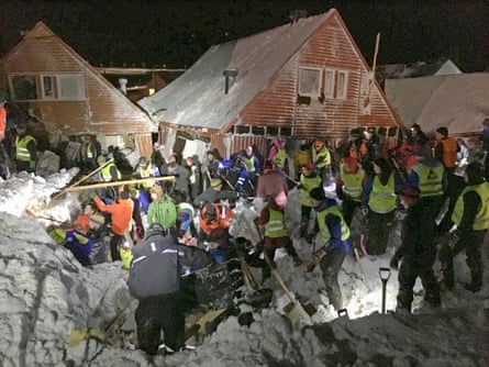 People digging into a pile of snow at night by several damaged houses