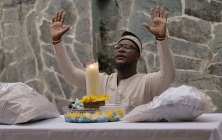A man dressed in white robes raises his hands in a blessing before a lit candle