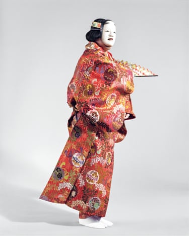 A performer in white face paint wears an ornate red floral kimono, holding a folded fan