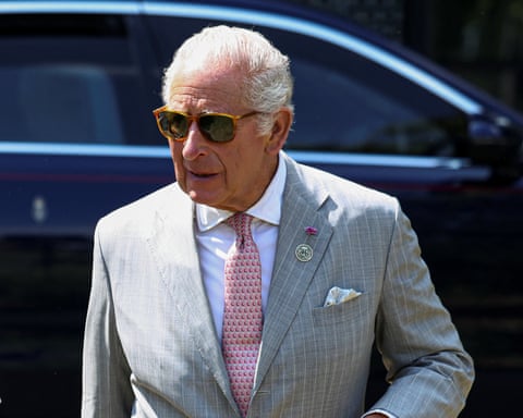 King Charles steps away from a dark car. He is wearing sunglasses and a pale grey suit with a red and silver tie and white shirt.