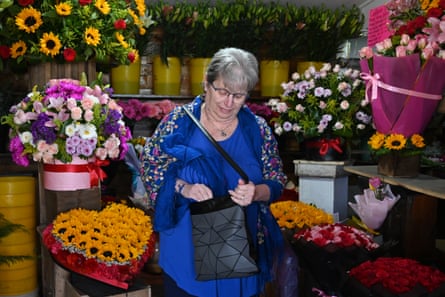 An older white woman dressed in bright blue looks into her purse among colorful flowers in buckets.