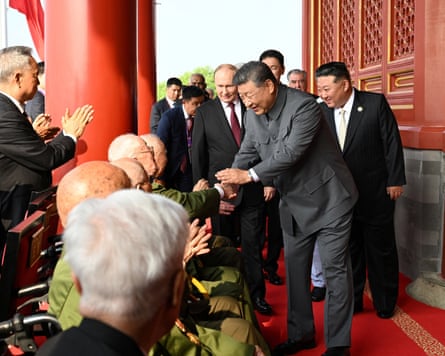 Xi Jinping shakes hands with an elderly man sat in a row with others, with Vladimir Putin and Kim Jong-un standing behind him