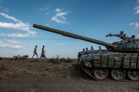 Two young men, one in a sarong and holding a stick and the other in tight trousers, walk past an abandoned tank behind which other young people are standing